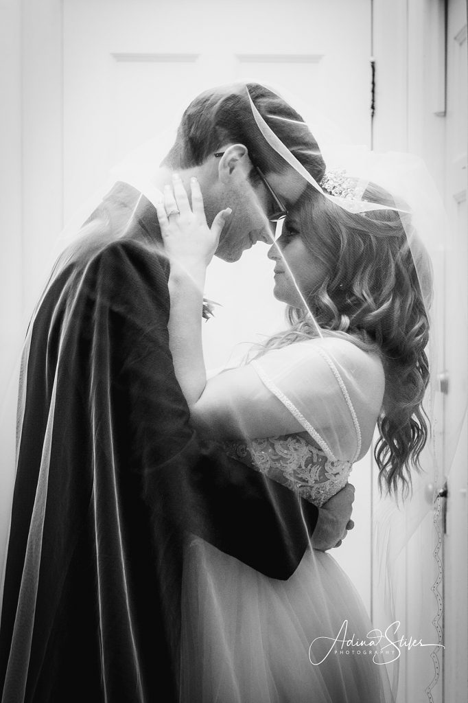 A groom stands under the veil and embraces his bride, while she places her hands on his face, and the moment is captured by Adina Stiles Photography.