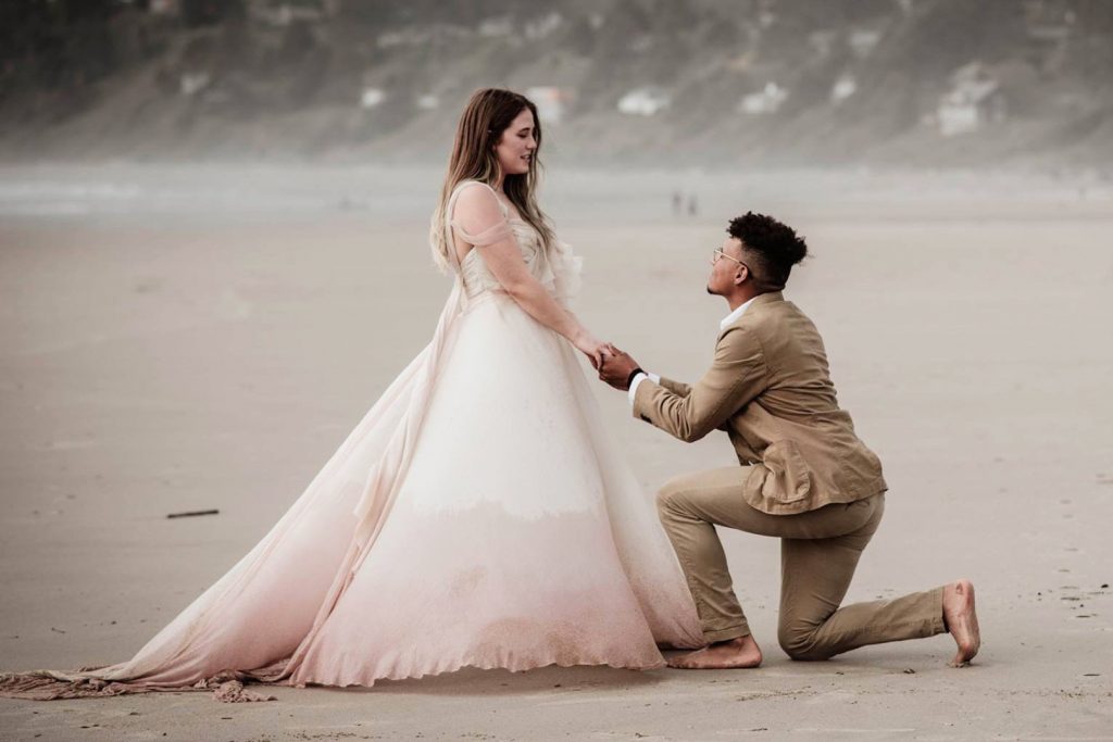 A man gets down on one knee while proposing on a holiday on the beach while his girlfriend wears a beautiful dress.