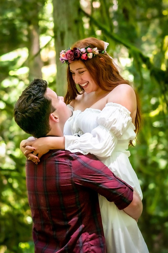 A woman with long red hair wearing a white dress and a flower crown looks down at her partner as he lifts her up in Grand Forest, Bainbridge Island, one of the best photo spots in Puget Sound.