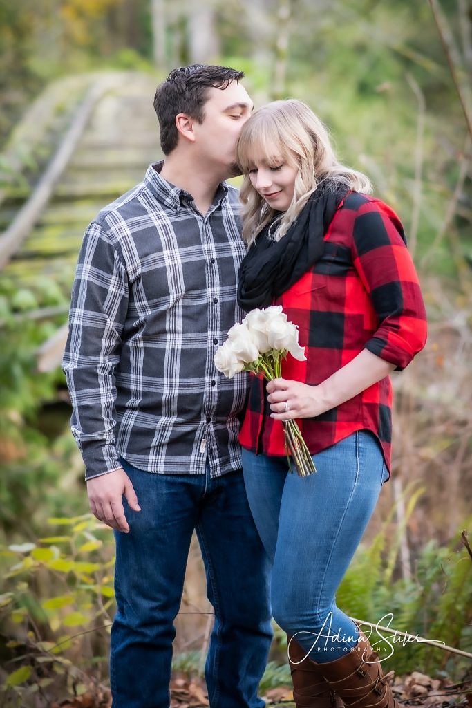 A man in a button-up shirt and woman in a red plaid shirt holding white flowers pose for engagement photos with Adina Stiles photography in Whatcom Falls Parks, Bellingham.