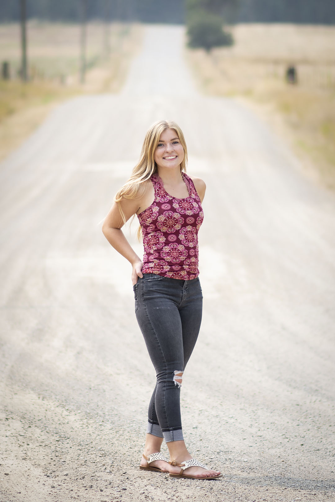 blonde-haired teenager stands on a dirt road smiling in this senior portraits shoot taken by adina stiles photography